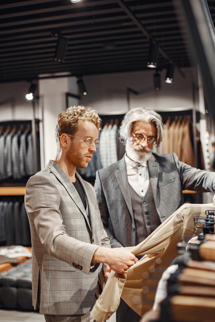 Two adult men examining suits in a stylish store with racks of clothing in the background.